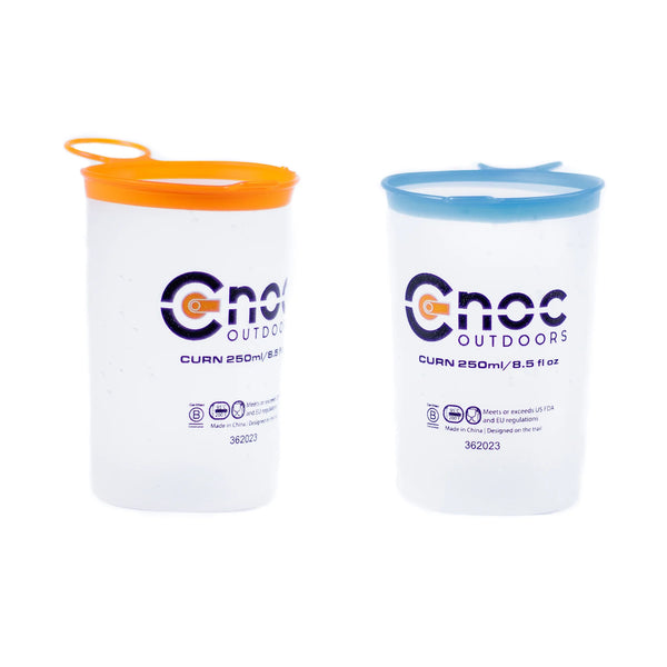 Two frosted Cnoc Outdoors CURN 250ml collapsible cups shown side by side on a white background, one with an orange silicone rim and one with a blue rim, both displaying product certification icons.