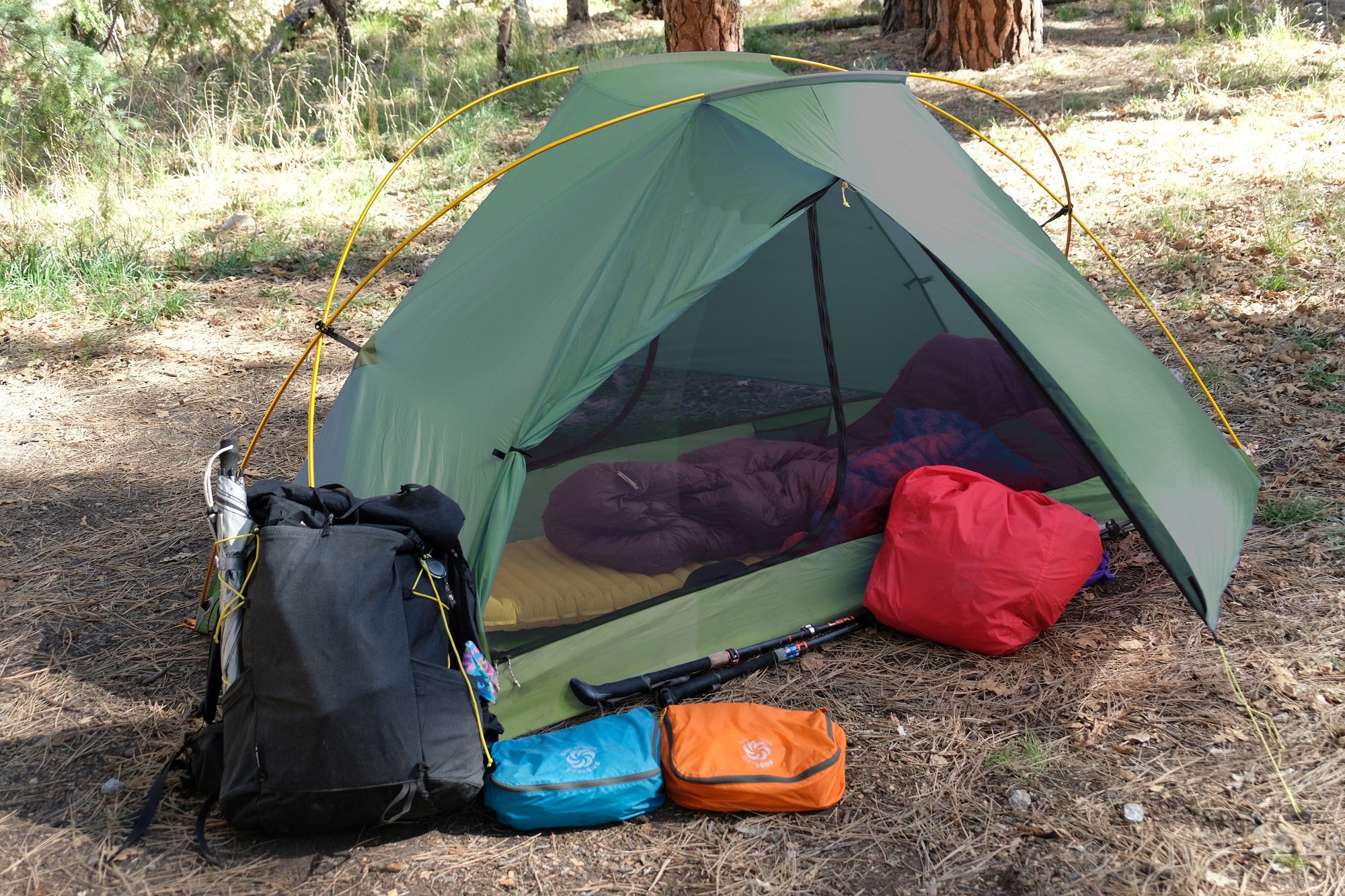 Lunar Orbiter at Camp with one vestibule door half open.  Swift X pack leaning up against tent with an umbrella in the side pocket and the red pack liner under the vestibule along with one blue and one orange packing pod and a couple of trekking poles laying down.  There is a sleeping bag and a sleeping pad inside the tent.  